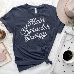 May include: A navy blue t-shirt with the text "Main Character Energy" in white cursive lettering. The shirt is laid out on a white surface with a book, a clock, a candle, a coffee cup, and a hat.