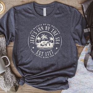 Puede incluir: Una camiseta azul oscuro con un diseño gráfico blanco de una casa de playa con palmeras y el texto "Jeff's Inn By The Sea Est 1717 You Gotta Ring The Bell!"