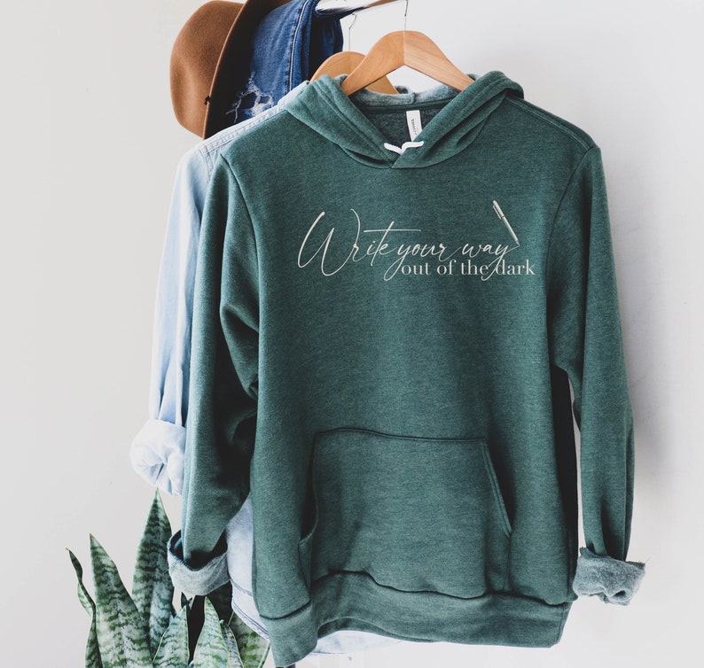 May include: A dark green hooded sweatshirt with the text "Write your way out of the dark" in white lettering. The sweatshirt has a pocket on the front.
