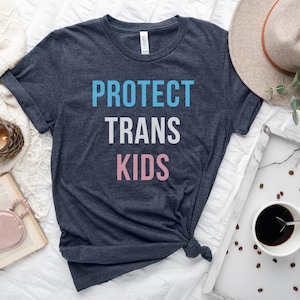 以下が含まれることがあります： 紺色のTシャツに「PROTECT TRANS KIDS」という文字が青、白、ピンクでプリントされています。