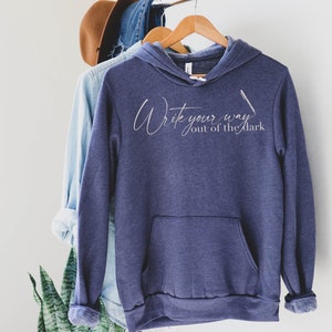 May include: A dark blue hooded sweatshirt with the text "Write your way out of the dark" in white script. A white pen is drawn next to the text.