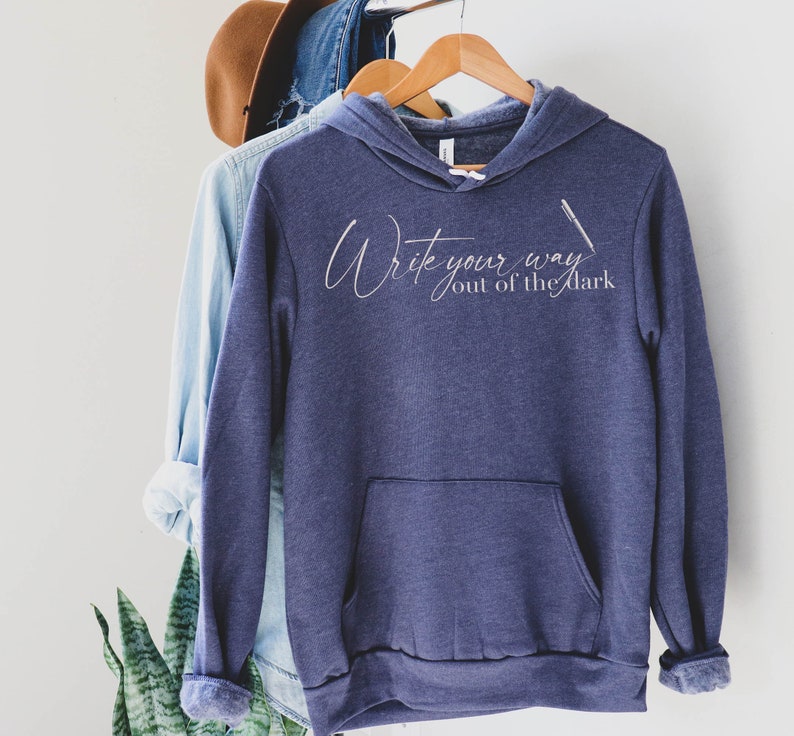 May include: A dark blue hooded sweatshirt with the text "Write your way out of the dark" in white script. A white pen is drawn next to the text.