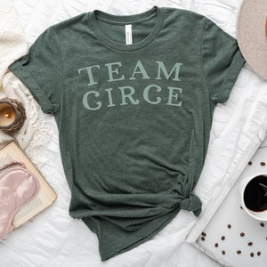 以下が含まれることがあります： 白い文字で「TEAM CIRCE」とプリントされた濃い緑色のTシャツ。Tシャツは裾で結ばれており、白い表面の上に置かれています。
