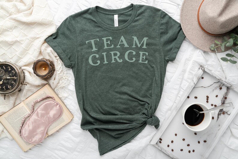 以下が含まれることがあります： 白い文字で「TEAM CIRCE」とプリントされた濃い緑色のTシャツ。Tシャツは裾で結ばれており、白い表面の上に置かれています。