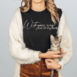 Könnte beinhalten: Eine Frau trägt ein schwarzes T-Shirt mit dem Text "Write your way out of the dark" in weißer Schrift. Sie hält ein kleines Tabby-Kätzchen in ihren Armen.
