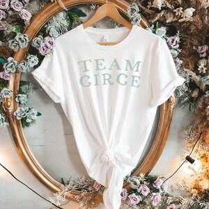 以下が含まれることがあります： 白地に薄い緑色の「TEAM CIRCE」の文字がプリントされたTシャツ。金色のハンガーにかけられており、白と茶色の花の装飾が背景にある。