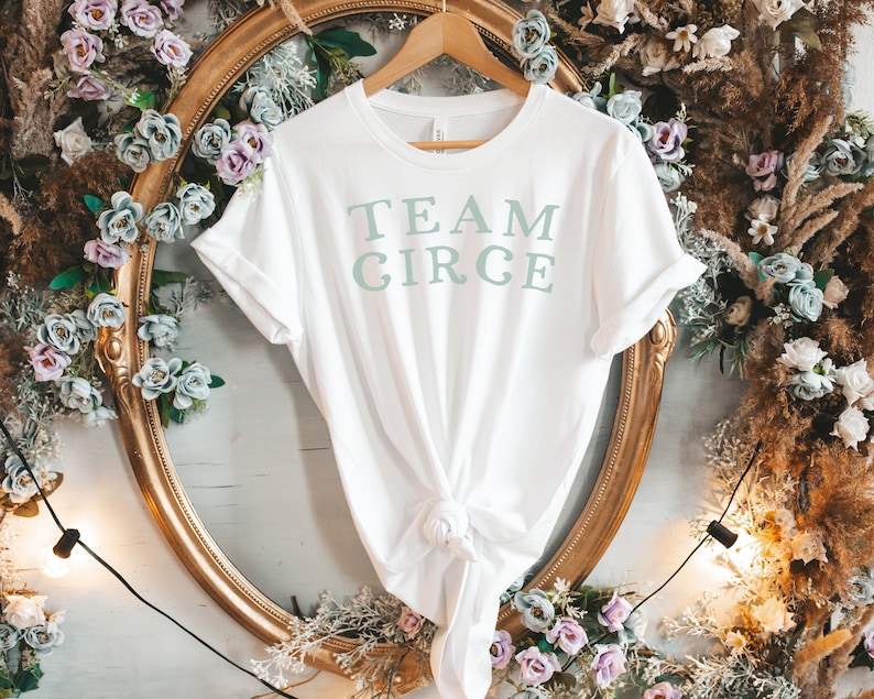 以下が含まれることがあります： 白地に薄い緑色の「TEAM CIRCE」の文字がプリントされたTシャツ。金色のハンガーにかけられており、白と茶色の花の装飾が背景にある。