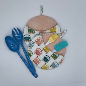 May include: Two blue kitchen utensils, a spoon and a fork, are placed on top of a white and colorful pot holder with a pattern of vintage vans. The pot holder is folded in half and has a light pink interior.