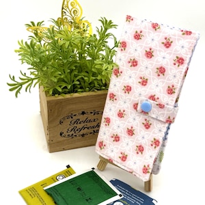 May include: A floral print fabric case with a blue button closure, displayed on a small wooden easel. The case features a pattern of pink roses on a white background. A wooden box with the words "Relax Refresh" is in the background.
