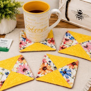 May include: A white mug with the word "Believe" in gold lettering sits on a floral coaster. Four additional floral coasters with yellow accents are arranged around the mug. A packet of Tazo tea and a decorative box are also visible.