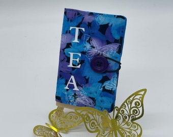 May include: A blue and purple fabric tea bag holder with a butterfly design and the word "TEA" printed in white. The holder has a purple button and a black elastic band.