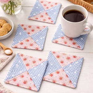 May include: Square fabric coasters with a patchwork design. Each coaster features a combination of pink floral fabric and blue gingham fabric. A white mug of coffee sits on one coaster. The coasters are part of a home decor theme.
