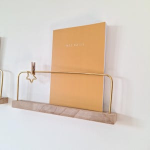 May include: A wall-mounted shelf with a wooden base and a gold-colored metal frame. A notebook with the text "MES NOTES" is held in place by a wooden clip and a star-shaped charm. The notebook is a warm, golden color.