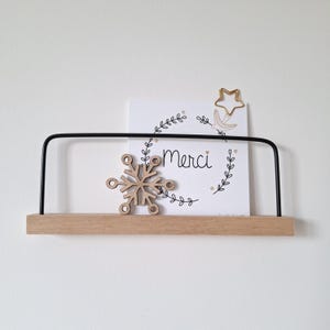 Peut inclure: Une étagère en bois avec une barre en métal noir, supportant une carte avec le mot "Merci" et un motif floral. Un flocon de neige en bois et un clip en forme d'étoile dorée sont également présents. L'étagère est fixée à un mur blanc.