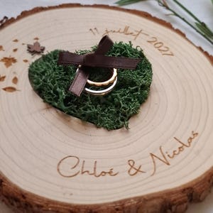 Peut inclure: Un porte-alliances en bois avec un cœur de mousse verte, accueillant deux alliances. Un ruban marron orne la mousse. Les noms "Chloé & Nicolas" et la date "11 juillet 2027" sont gravés sur le bois.