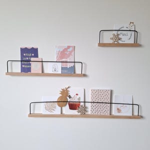 Può includere: Tre mensole da parete in legno con ringhiere in metallo nero, che espongono varie carte e oggetti decorativi. Le carte presentano testo e illustrazioni, tra cui un ananas e un cupcake. Le mensole sono montate su una parete bianca.