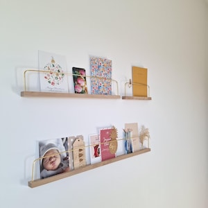 Op de afbeelding: Houten wandplanken met messingkleurige metalen relingen, met diverse kaarten, foto's en een telefoon. De planken zijn aan een witte muur bevestigd en tonen een minimalistisch ontwerp. De kaarten bevatten een kerstgroet en een babyfoto.