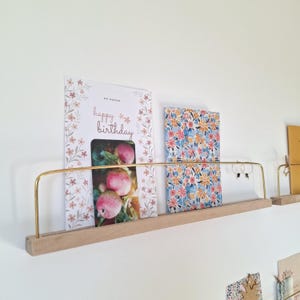 Może przedstawiać: Drewniana półka z mosiężną szyną prezentuje kartkę urodzinową z napisem "happy birthday" i kwiatowym wzorem. Wyświetlana jest również druga karta z kwiatowym wzorem. Półka jest zamontowana na białej ścianie.