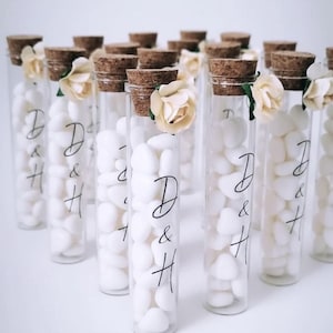 Op de afbeelding: Transparante glazen buisjes gevuld met witte stenen, afgedekt met kurken en kleine crèmekleurige bloemen. Elke buis heeft de letters "D & H" in zwart gedrukt. Een decoratief huwelijksbedankje.