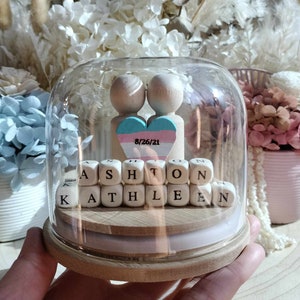 May include: A glass dome with a wooden base and a small wooden heart with the date 8/26/21. The heart is painted in blue, white, and pink colours. The dome also contains wooden blocks spelling out the names Ashton and Kathleen.