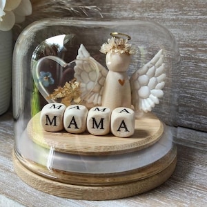 Pode incluir: Uma figura decorativa de anjo dentro de uma cúpula de vidro. O anjo tem asas brancas, uma coroa floral e um coração dourado. A palavra "MAMA" é escrita com blocos de letras de madeira. Uma peça de resina em forma de coração com flores também está dentro.