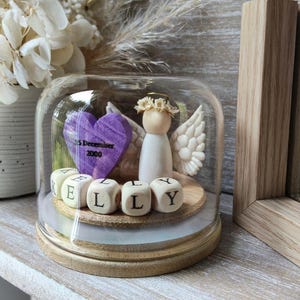 May include: A glass dome on a wooden base contains a wooden angel figurine with wings and a floral crown. A purple heart with the date "5 December 2000" and wooden letter blocks spelling "KELLY" are also inside the dome.