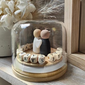 Op de afbeelding: Een decoratieve glazen stolp met een houten basis herbergt twee houten poppetjes, één wit met een hart en de andere zwart. Houten blokken spellen "WAYS" en "3 YRS". Een romantisch aandenken.
