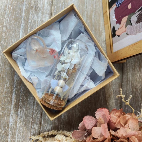 Könnte beinhalten: Eine dekorative Geschenkbox mit einer Glasflasche mit weißen Blumen und Holzperlen. Ein herzförmiges Harzstück befindet sich ebenfalls in der Box, die mit Seidenpapier ausgekleidet ist. Die Box steht auf einer Holzoberfläche.