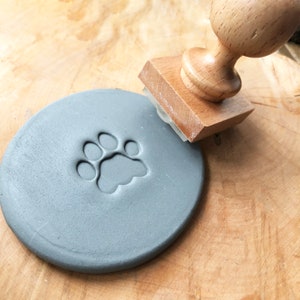 Op de afbeelding: Een houten stempel met een pootprintontwerp wordt gebruikt om een afdruk op een grijs kleioppervlak te maken.