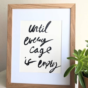 May include: A framed print with the text "Until every cage is empty" in black calligraphy on a white background. The frame is made of light brown wood.