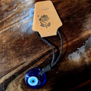 May include: A blue evil eye charm necklace with a black leather cord. The charm is round and has a white eye with a blue pupil.