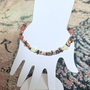 May include: A beaded bracelet featuring a mix of orange, brown, and gold-colored beads. The bracelet includes small square beads with etched symbols and is displayed on a white hand-shaped stand.
