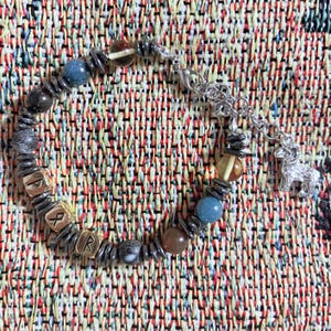 May include: A beaded bracelet with a silver clasp and a charm of a ram. The bracelet features a mix of dark gray, gold, and colorful beads, including blue, brown, and clear stones. Gold-colored square beads spell out the word "Rune".