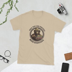 Pode incluir: Camiseta bege com um gráfico circular com um urso usando um chapéu de guarda florestal, segurando livros e apontando. O texto diz "ONLY YOU CAN PREVENT BOOK BURNINGS". Itens adicionais incluem uma câmera, óculos, uma carteira e um telefone.