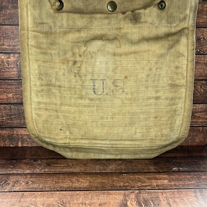 May include: A tan canvas bag with a flap and metal snaps. The bag has a U.S. marking on the front.
