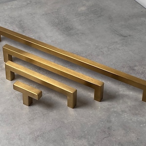 May include: Five gold-toned metal cabinet pulls in various sizes. The pulls are rectangular with rounded edges. The smallest pull is a T-shape.