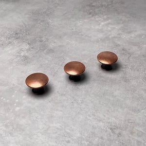 May include: Three round, copper-coloured cabinet knobs with a slightly textured finish.