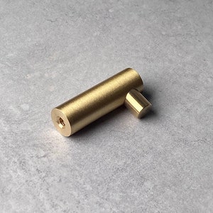 May include: A brushed gold metal hook with a cylindrical shape and a rounded end. The hook is designed for hanging items on a wall.