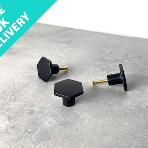 May include: Three black hexagonal cabinet knobs with gold screws. The knobs are on a grey surface.