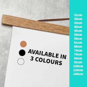 May include: A white poster with the text "AVAILABLE IN 3 COLOURS" in black. Three circles are shown below the text: a brown circle, a black circle, and a white circle. A wooden frame with a brown leather strap is at the top of the poster. A list of sizes in centimetres is shown on the right side of the poster.