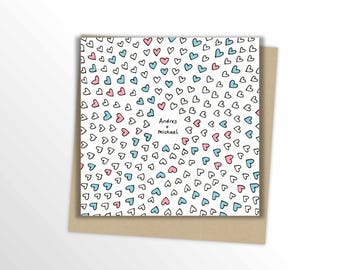 May include: A square greeting card with a white background, covered in small, outlined hearts in black, pink, and light blue. The card includes the names "Andres + Michael" in a handwritten font. The card is placed on a brown envelope.