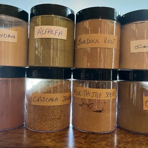 May include: A collection of clear plastic jars with black lids, each labeled with handwritten names of herbs and supplements. The jars contain various dried herbs and powders, including Ashwagandha, Alfalfa, and Milk Thistle Seed.