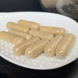 May include: A white ceramic spoon with a textured surface holds a group of light brown capsules. The capsules are arranged in a semi-circle shape.
