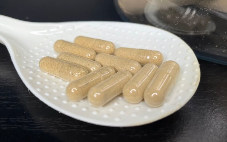 May include: A white ceramic spoon with a textured surface holds a group of light brown capsules. The capsules are arranged in a semi-circle shape.