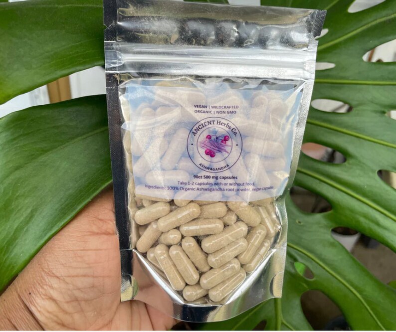 May include: A silver pouch with a clear plastic window showing brown capsules. The pouch is labeled "ANCIENT Herbs Co. Ashwagandha 90ct 500 mg capsules Vegan | Wildcrafted Organic | Non GMO Take 1-2 capsules with or without food 100% Organic Ashwagandha root powder, vegan capsules"