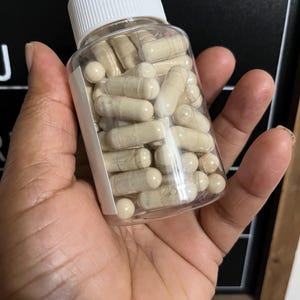 May include: A clear plastic bottle filled with beige capsules, with a white lid. The bottle is being held in a person's hand.