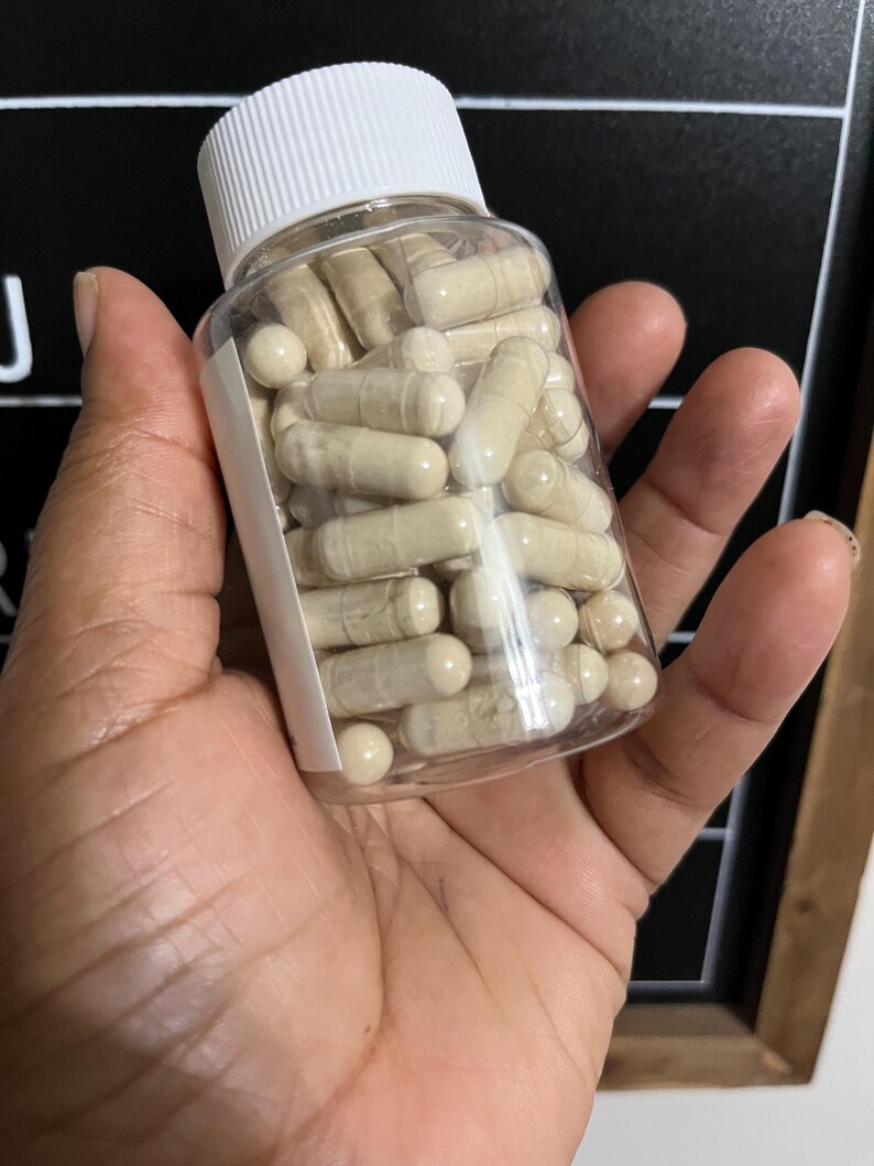 May include: A clear plastic bottle filled with beige capsules, with a white lid. The bottle is being held in a person's hand.