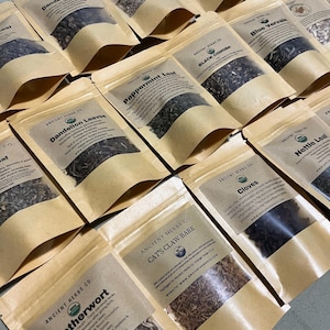 May include: A collection of brown paper bags filled with dried herbs and labeled with the name of the herb and the company name "Ancient Herbs Co.".