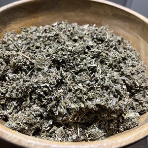 May include: A wooden bowl filled with dried, crumbled green leaves. The leaves have a muted green and brown color, with some lighter, almost white, pieces mixed in. The bowl itself is made of light-colored wood.