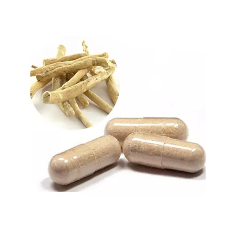 May include: Three capsules of a light brown color, with a clear outer shell. The capsules are arranged in a row on a white surface. A pile of dried, light brown, root-like pieces are shown in the background.